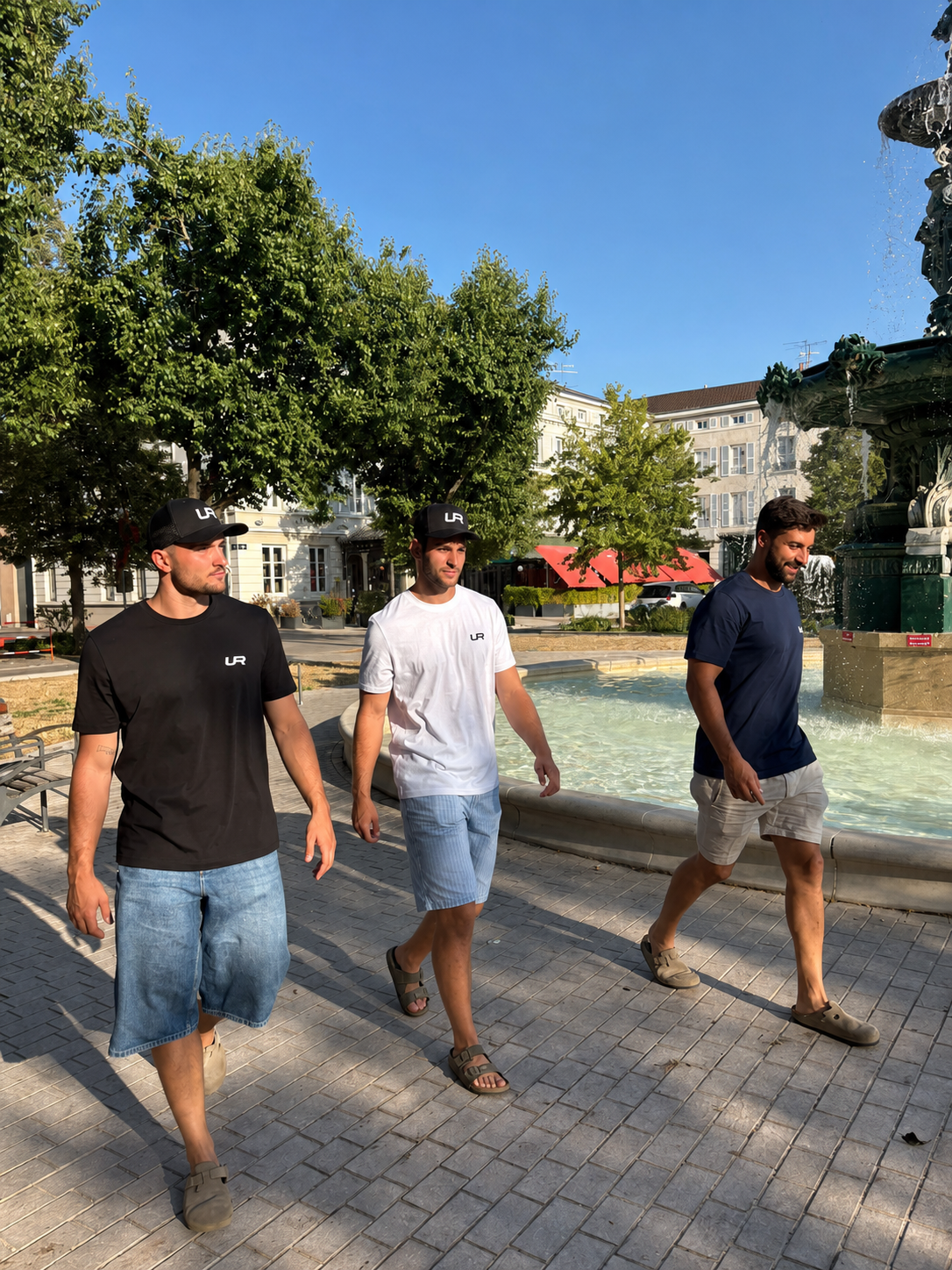 Trois hommes portant des vêtements URGEZA devant une fontaine en plein air